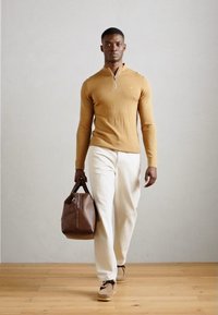 Maglione con zip color cammello abbinato a pantaloni bianchi e borsa in pelle marrone. Il modello è in piedi su un pavimento in legno contro un muro neutro.