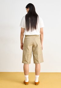 Beige tailored shorts made of cotton blend, knee-length, with back pockets, paired with a white shirt and orange slip-on shoes.
