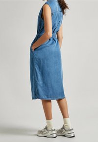 Denimklänning i ljusblått, utan ärmar med knapprad framtill, åtsittande midja och sidofickor. Bärs med grå och vita sneakers.