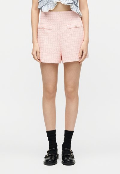 Γυναίκα που φοράει υφασμένα ροζ ανοιχτού χρώματος σορτς με ψηλή μέση και κουμπιά, σε συνδυασμό με μαύρες κάλτσες και μαύρα παπούτσια με αγκράφα, στέκεται μπροστά σε λευκό φόντο.