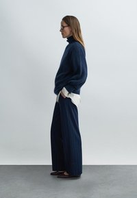 Marineblauer Strick-Rollkragenpullover mit lockerem Schnitt, über einem weißen Hemd getragen. Kombiniert mit weitem, gestreiftem Hosenrock und dunklen flachen Schuhen.