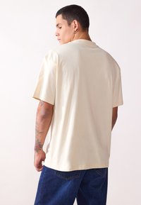 Camiseta de algodón beige con un corte relajado, de manga corta y cuello redondo, mostrada desde la parte de atrás, combinada con jeans azules.