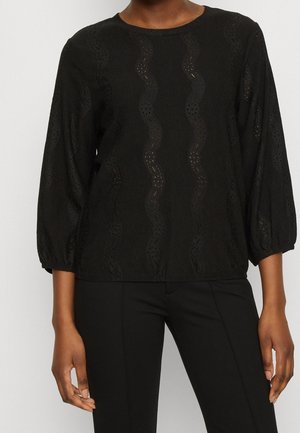 Blouse noire à manches longues avec motifs en dentelle ondulés, associée à un pantalon noir ajusté, montrée du cou aux hanches sur une personne debout.