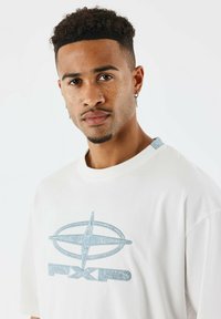 Camiseta de algodón blanca con un logo y diseño azul en el frente, cuello redondo y detalles de mezclilla en los hombros.