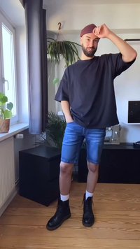 T-shirt oversized preta, calções de ganga azuis, meias brancas, botins pretos. Ambiente interior com plantas e uma máquina de costura ao fundo.