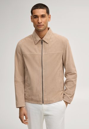 Mann trägt eine hellbraune Jacke mit Reißverschluss und weiße Hose und steht vor einem einfarbigen Hintergrund, eine Hand in der Tasche.