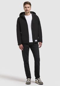 Sort letvægtsjakke med hætte, frontlynlås og sidelommer, parret med en hvid t-shirt og mørke jeans, sat mod en neutral baggrund.