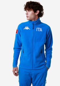 Giacca in fleece blu con colletto alto, due tasche con zip e logo Kappa. Presenta la bandiera italiana sulla manica e la scritta in grassetto "ITA".