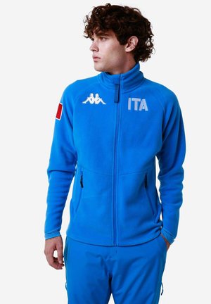 Giacca in fleece blu con colletto alto, due tasche con zip e logo Kappa. Presenta la bandiera italiana sulla manica e la scritta in grassetto "ITA".