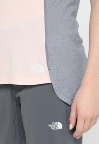 Jasnoróżowy i niebieski sportowy top ze wzorem, z rozcięciem po boku, z małym białym logo na dolnym brzegu. W połączeniu z szarymi spodniami.