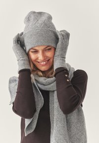 Grijze gebreide muts met een gestructureerd patroon, bijpassende grijze gebreide handschoenen en een grijze sjaal gedrapeerd over een donker shirt.