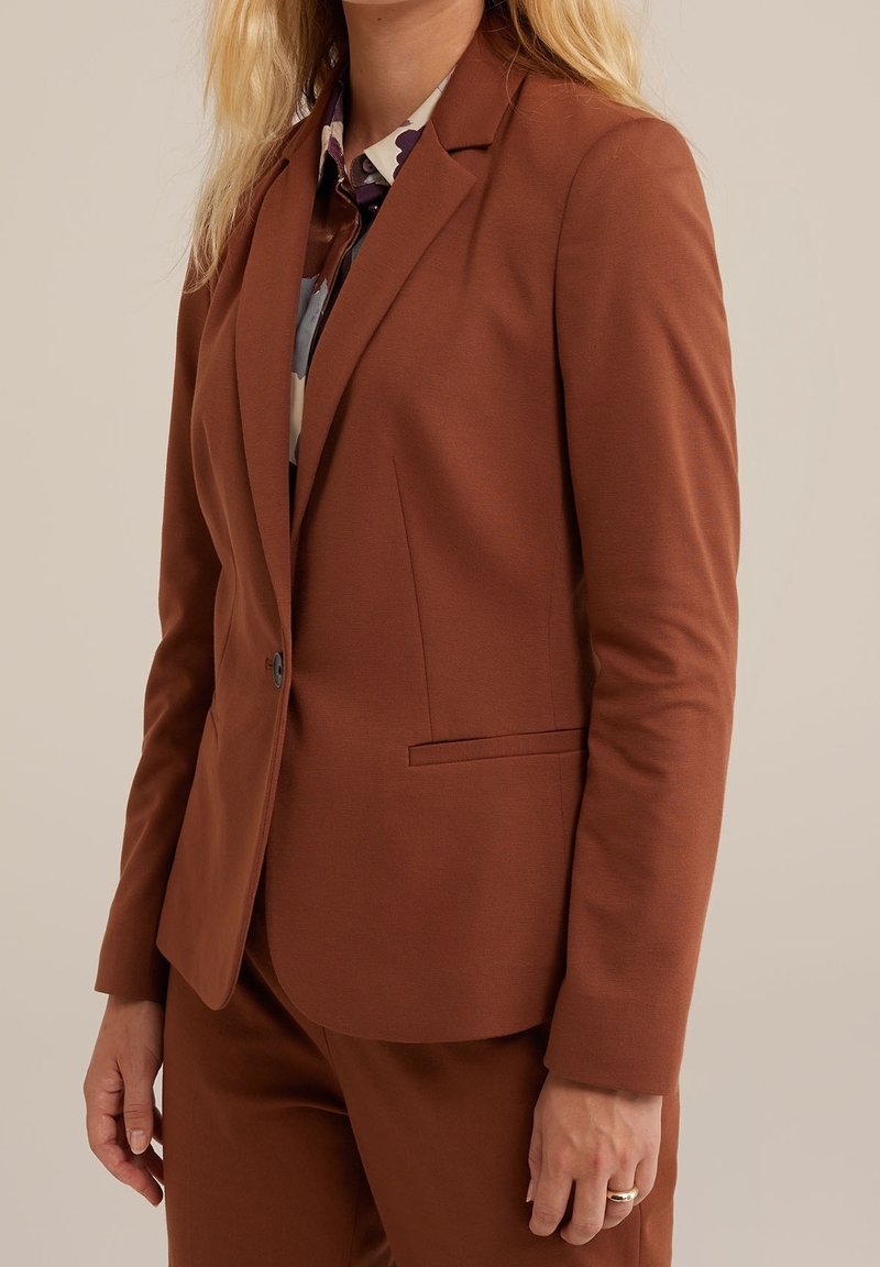 Brun skräddarsydd kavaj med slag i halsen, enkel knappstängning och sidofickor, tillverkad av ett slätt tyg. Bärs över en mönstrad skjorta.