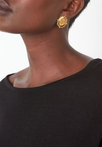 Boucles d'oreilles en métal doré avec un design rond en relief, ornées d'accents en forme d'éclats de soleil et d'une finition texturée, portées sur un haut foncé.