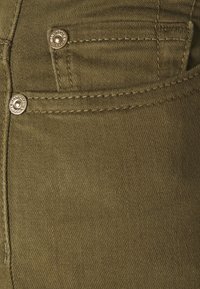 Tela de mezclilla verde oliva con detalle de bolsillo frontal, que cuenta con herrajes de botón plateado y una sutil textura de costura.