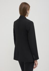 Blazer nero, design sartoriale, maniche lunghe, colletto a punte, chiusura con un solo bottone e tre bottoni decorativi su ciascuna manica. Tessuto di texture liscia.