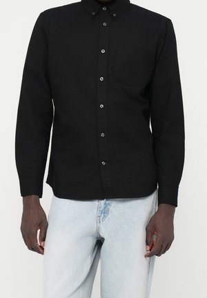 Mann trägt ein schwarzes, langärmliges Hemd mit Knöpfen und Kragen sowie hellblaue Jeans, steht vor einem schlichten weißen Hintergrund.