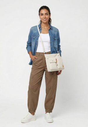 Woman wearing a denim shirt, white tank top, brown cargo pants, white sneakers, and a beige crossbody bag, standing and smiling.