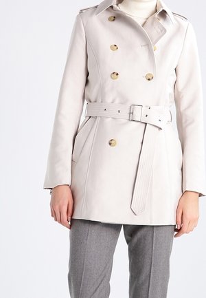 Vrouw draagt een beige dubbelgeknoopte trenchcoat met bruine knopen en riem, gecombineerd met grijze broek en een witte coltrui.