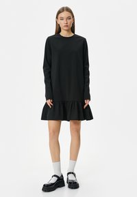 Musta pitkähihainen mekko, jossa on röyhelöhelma, valmistettu sileästä kankaasta. Mallilla on jalassaan paksut mustat kengät ja valkoiset sukat. Yksinkertainen, minimalistinen muotoilu.