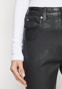 Μαύρα τζιν παντελόνια από υλικό ντενίμ με υφή, διαθέτουν πέντε τσέπες και ασημί λεπτομέρειες. Το χέρι resting στον πλευρικό ραφά.
