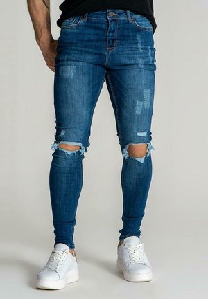 Uske traperice u srednje plavom denimu s izrezima na koljenima; sadrži klasičan dizajn s pet džepova i uski kroj, uparen s bijelim tenisicama.