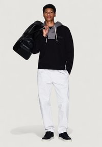 Joven en sudadera negra superpuesta a un suéter gris, pantalones blancos, zapatillas negras, llevando una bolsa de deporte negra al hombro, de pie contra un fondo blanco.