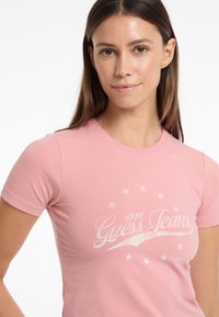Šviesiai rožinė medvilninė marškinėliai su trumpomis rankovėmis, apvaliu kaklu ir balta grafika su užrašu "Guess Jeans" ir žvaigždėmis.
