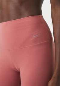 Rózsaszín sportleggings, magas derékkal, sima textúrával és egy kis szürke Nike logóval az oldalon.