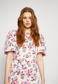 Vestido floral com mangas curtas e volumosas, em cores pastel incluindo rosa e azul, e um fundo bege claro com padrões de flores diversas.