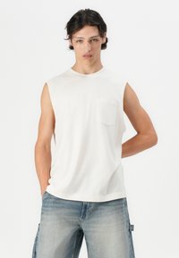 Αμάνικη λευκή βαμβακερή μπλούζα με στρογγυλή λαιμόκοψη και μικρή τσέπη στο στήθος, συνδυασμένη με ανοιχτό μπλε τζιν σορτς.