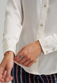 Hvit langermet skjorte med trykknapper, med broderte mansjetter. Mansjettene har "Elaine" og "Mona" i lys tråd mot stoffet.