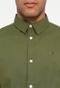 Zāles zaļš krekls ar pogājamu apkakli un caurspīdīgām pogām. Piedāvā nelielu logotipu ar sarkaniem, baltajiem un ziliem akcentiem uz krūšu daļas.