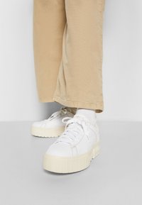 Personne portant un pantalon beige, des chaussettes blanches et des baskets à plateformes blanches avec des semelles épaisses et texturées, debout sur un sol gris.