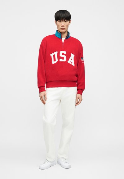 Κόκκινο φλις πουλόβερ με λευκές επιστολές «USA», γιακά με φερμουάρ και πράσινες λεπτομέρειες. Συνδυάζεται με λευκό παντελόνι και λευκά αθλητικά παπούτσια.