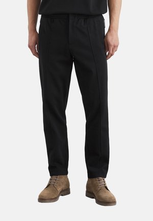 Schwarze Hose mit elastischem Bund, vertikalen Nähten und lockerem Schnitt. Das schmal zulaufende BeinDesign ergänzt braune Wildleder-Schnürstiefel.