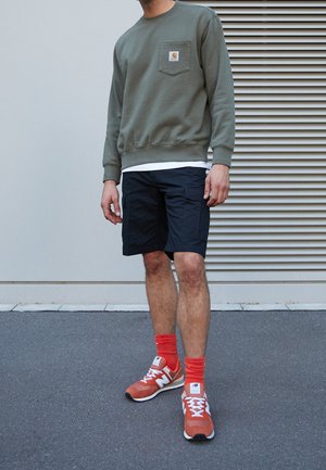 Mężczyzna w oliwkowej bluzie, czarnych szortach typu cargo, jasnopomarańczowych skarpetkach oraz pomarańczowo-białych tenisówkach stoi na chodniku.