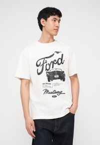 Νεαρός άνδρας με λευκό μπλουζάκι που απεικονίζει το μαύρο λογότυπο Ford Mustang, εικονογράφηση του αυτοκινήτου Mustang 1964, και σκούρα τζιν, όρθιος με το ένα χέρι στην τσέπη.