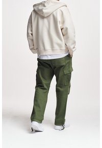 Felpa con cappuccio beige dal fit rilassato, abbinata a pantaloni cargo verdi larghi con tasche laterali e sneakers bianche.