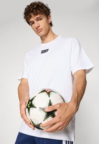 Hvit bomull T-skjorte med en liten svart og rød logo på brystet. Personen holder en hvit fotball med svarte og rosa detaljer.
