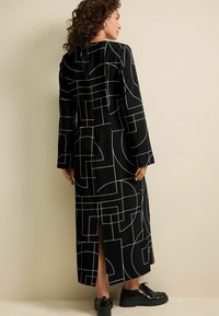 Mustapuku pitkä mekko, jossa on valkoinen geometrinen kuvio, pitkät hihat ja sivuhalkiot, yhdistettynä mustiin kenkiin. Teksturoitu kangas.