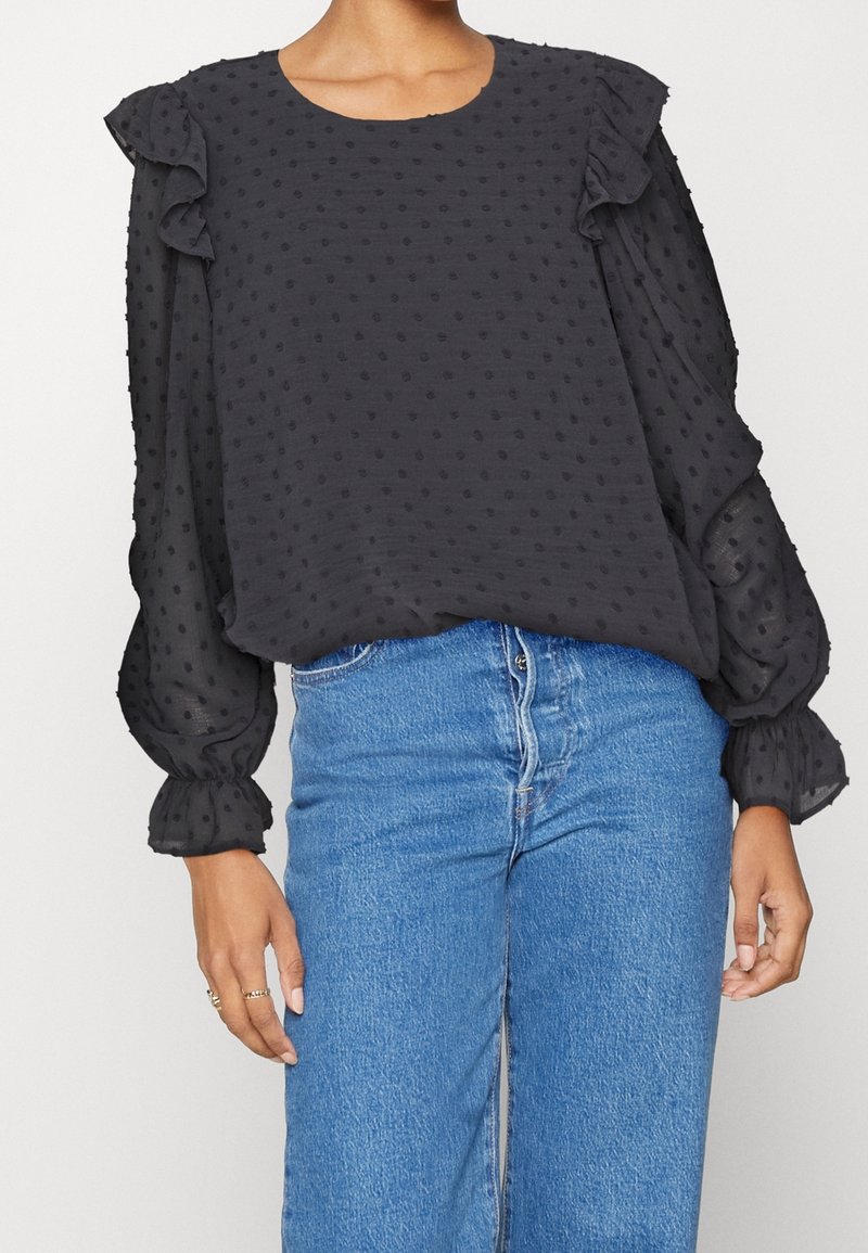 Blouse noire transparente avec des pois texturés, épaules et poignets à volants, portée avec un jean en denim bleu taille haute.