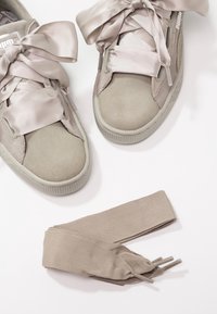 Paire de baskets beiges Puma avec de larges lacets en satin noués en nœuds, accompagnée d'une paire supplémentaire de lacets plats beiges sur une surface blanche.