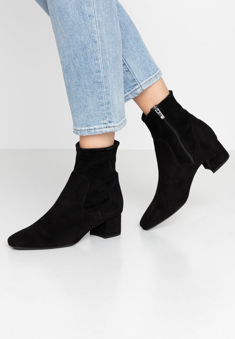 Personne portant un jean bleu clair et des bottines en daim noir avec des talons blocs et des fermetures éclair latérales sur un sol blanc.