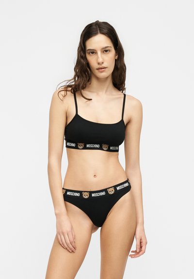 Femme en ensemble de lingerie noir deux pièces avec logo Moschino et imprimés d'oursons sur les bandes élastiques, debout devant un fond clair uni.