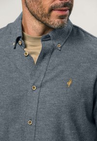 Szürke gombos ing, amely texturált anyagból készült és gallérral rendelkezik, kis arany logóval és világosbarna gombokkal. Bézs aláöltözet fölött viselik.