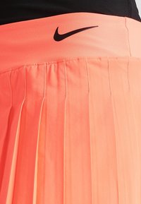 Gonna sportiva plissettata in un vivace tessuto arancione neon, presenta una vita aderente con il logo Nike nero. La texture è liscia con pieghe verticali.