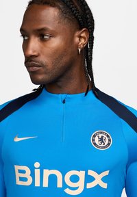Maglietta sportiva blu con colletto alto, con accenti blu navy, logo del Chelsea FC e uno swoosh Nike oro. Logo "Bingx" in beige chiaro.