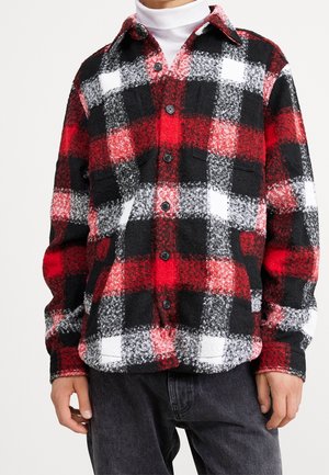 Man draagt een rood, zwart en wit geruit overhemd van flanel met knoopsluiting, twee borstzakken en een donkere broek.