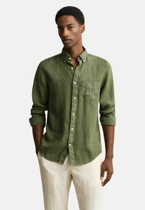 Young man wearing a green buttoned linen shirt with rolled-up sleeves and light beige striped pants, standing with hands in pockets.