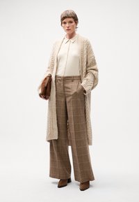 Beige gebreide cardigan over een crème blouse en bruine broek met ruitpatroon en wijde pijpen, gecombineerd met bruine enkellaarzen en een kleine tas.